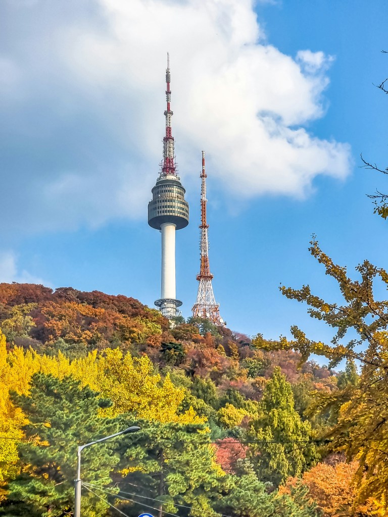 Namsan Tower rising above colorful autumn foliage, with views of the Seoul skyline in the background.