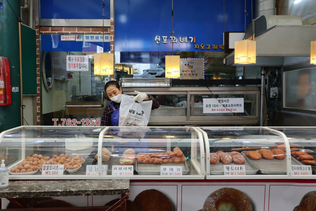 A Korean street vendor displaying freshly fried kkwabaegi (twisted donuts) and red bean chapssal donuts.